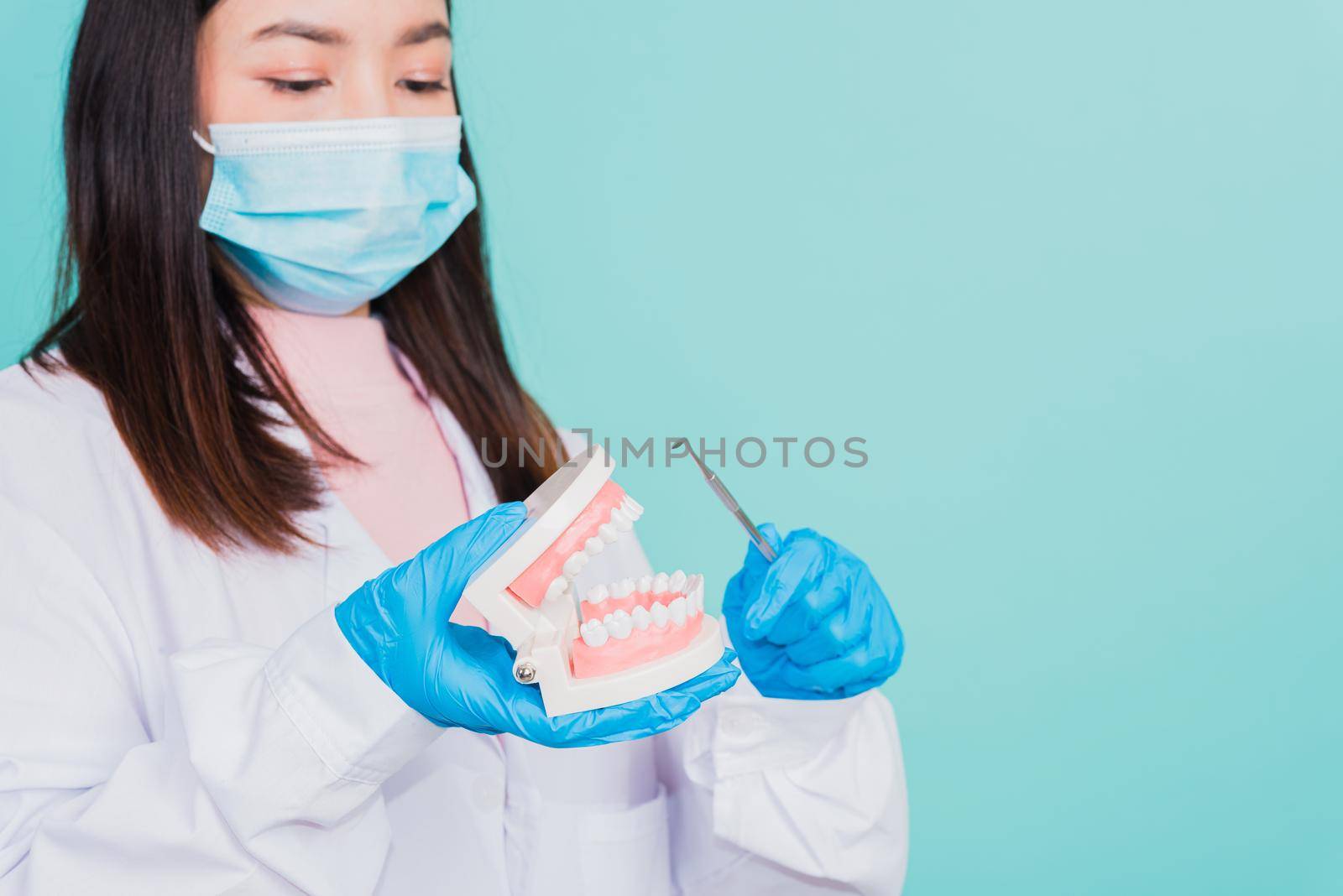 Asian beautiful woman dentist holding professional tool and pointing model teeth denture, female doctor checking denture isolated on blue background. Dental hygiene surgery health care concept