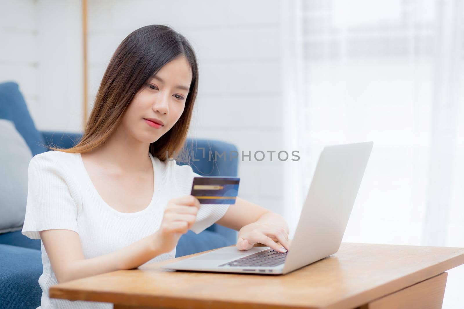 Young asian woman sit on sofa using laptop computer shopping online with credit card buying to internet, happy girl payment with e-business on couch, purchase and payment, business concept.