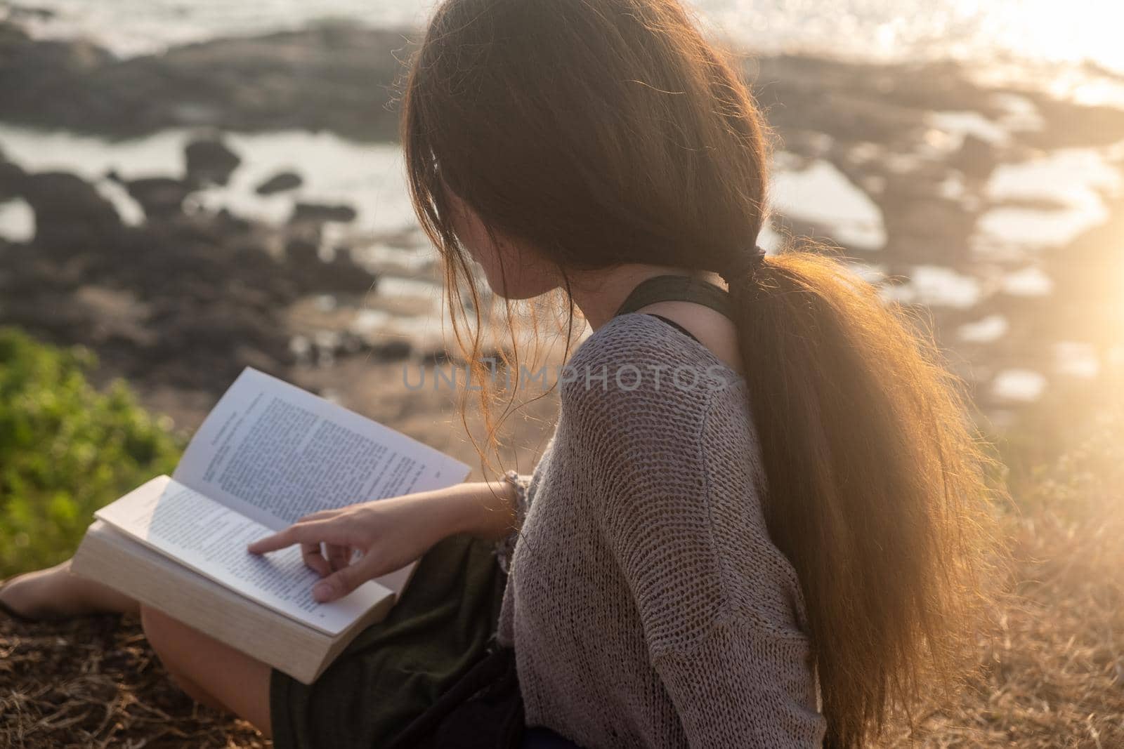 Attractive Asian woman is sitting on the edge of the mountain with a sea view, reading a book. From the back view. Sunlight