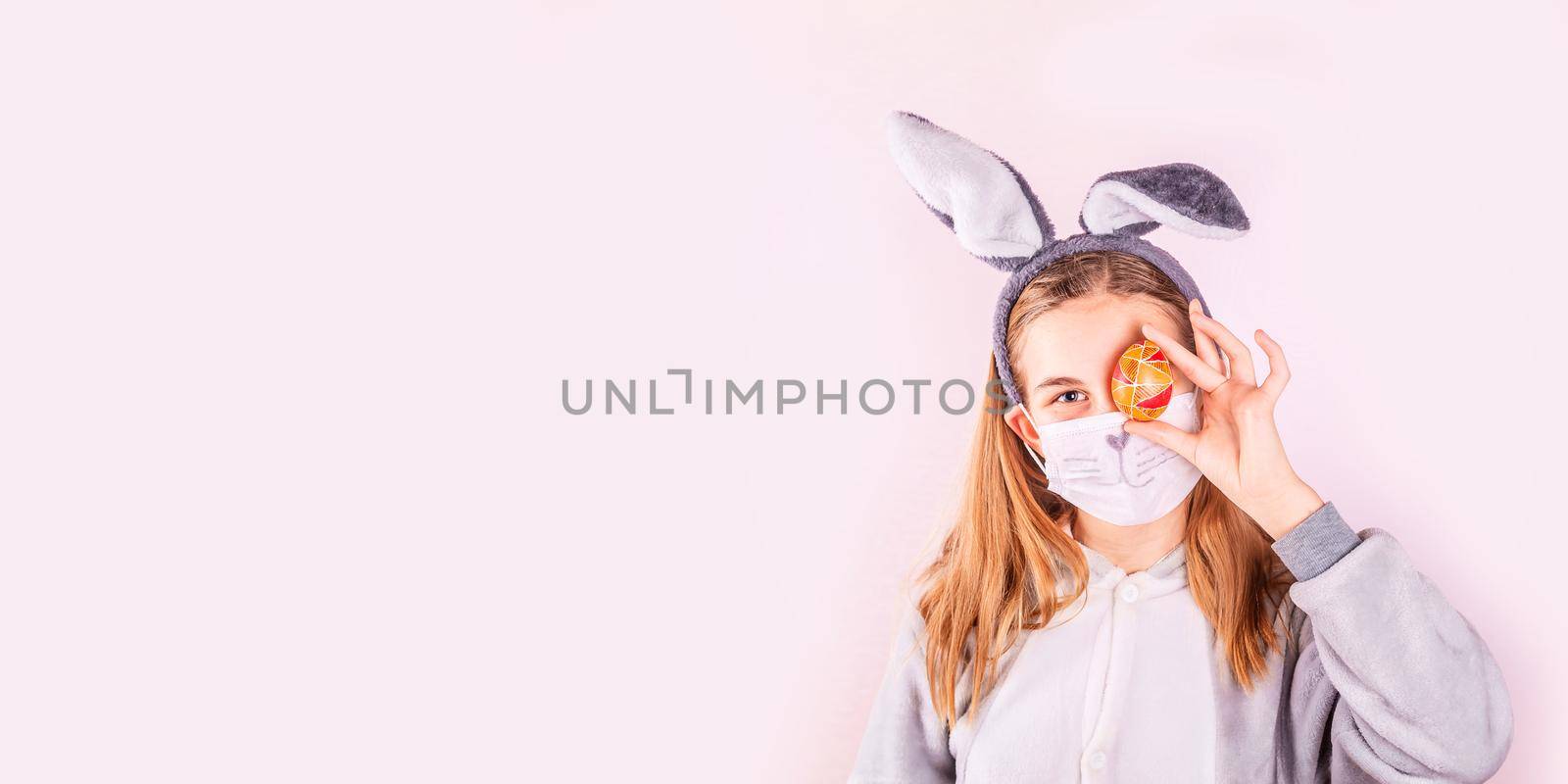 Girl in rabbit bunny ears on head and protective mask with colored eggs on pink background. Cheerful smiling happy child. Covid Easter holiday banner