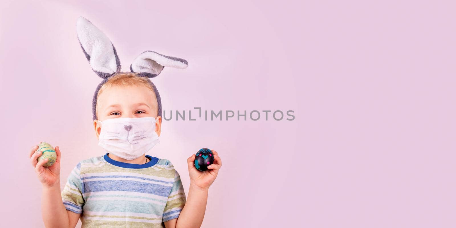 Cute baby boy in rabbit bunny ears on head and protective mask with colored eggs on pink background. Cheerful smiling happy child. Covid Easter holiday banner