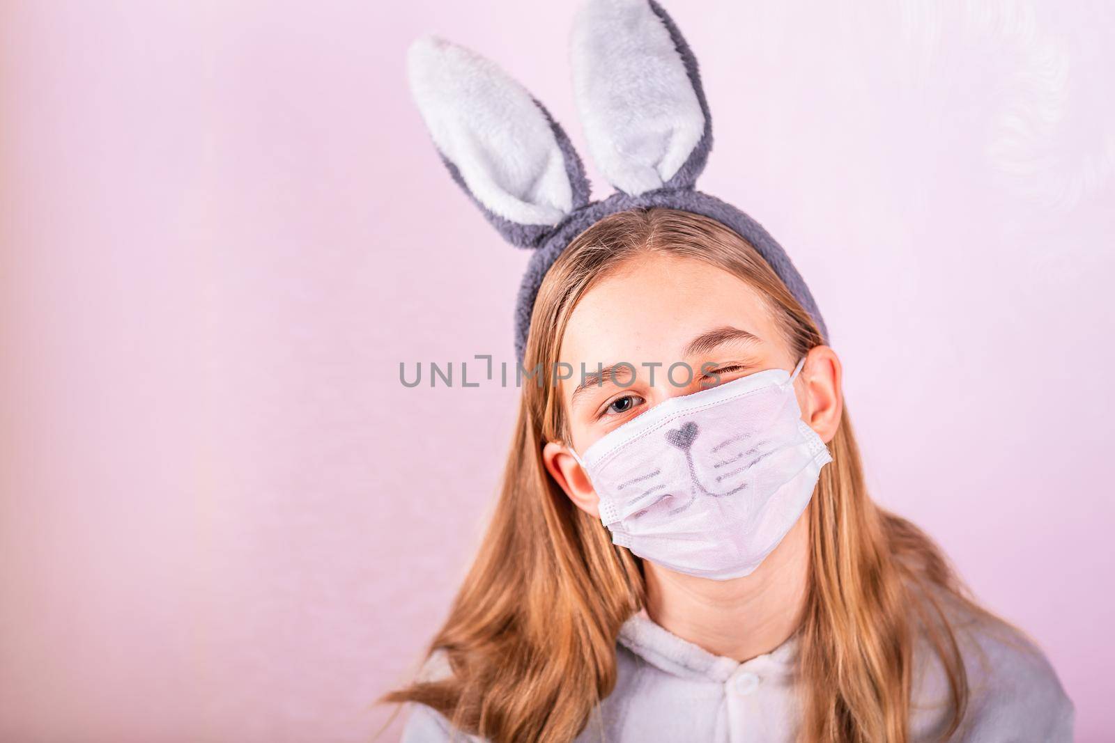 Girl in rabbit bunny ears on head and protective mask with colored eggs on pink background. Cheerful smiling happy child. Covid Easter holiday