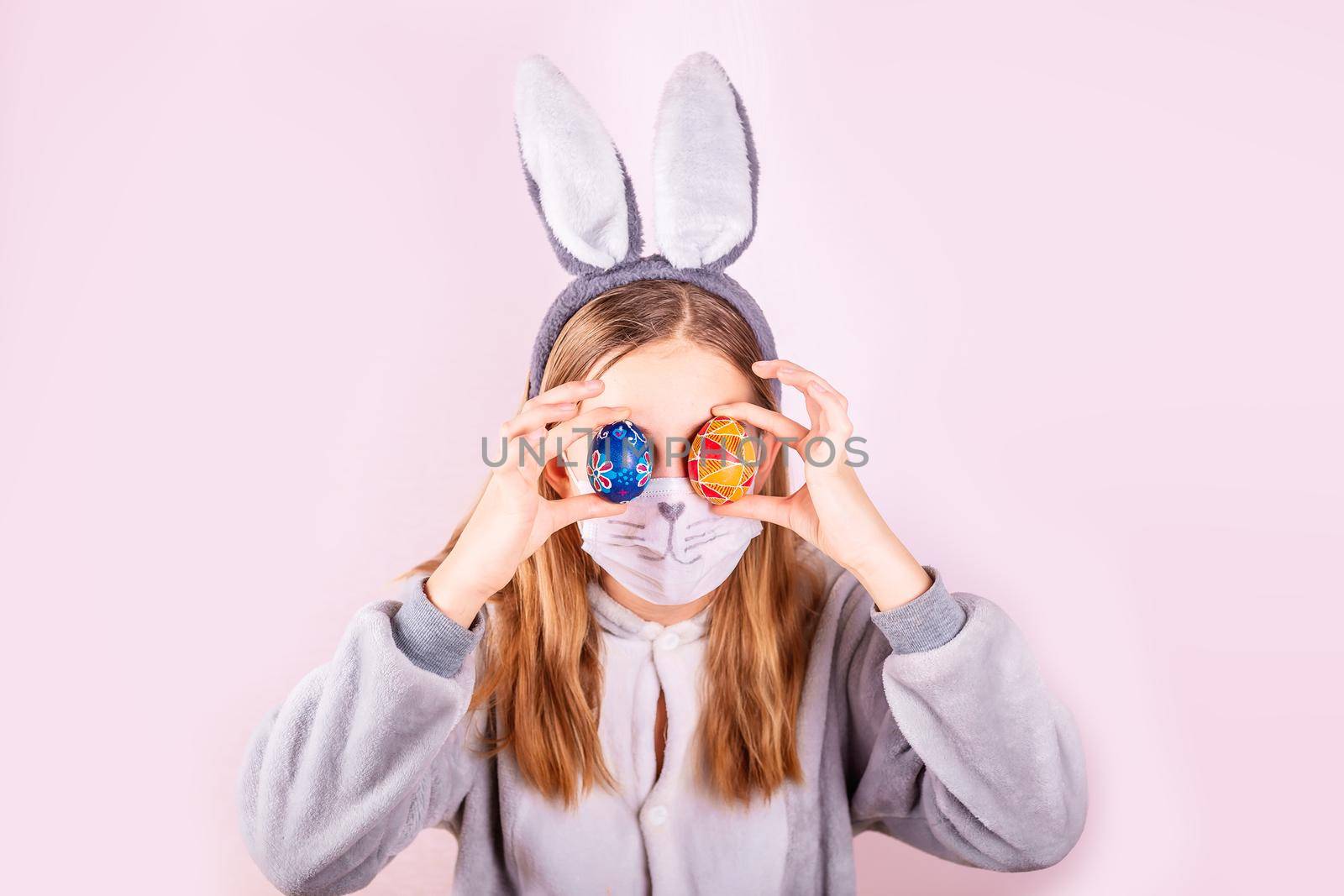 Girl in rabbit bunny ears on head and protective mask with colored eggs on pink background. Cheerful smiling happy child. Covid Easter holiday