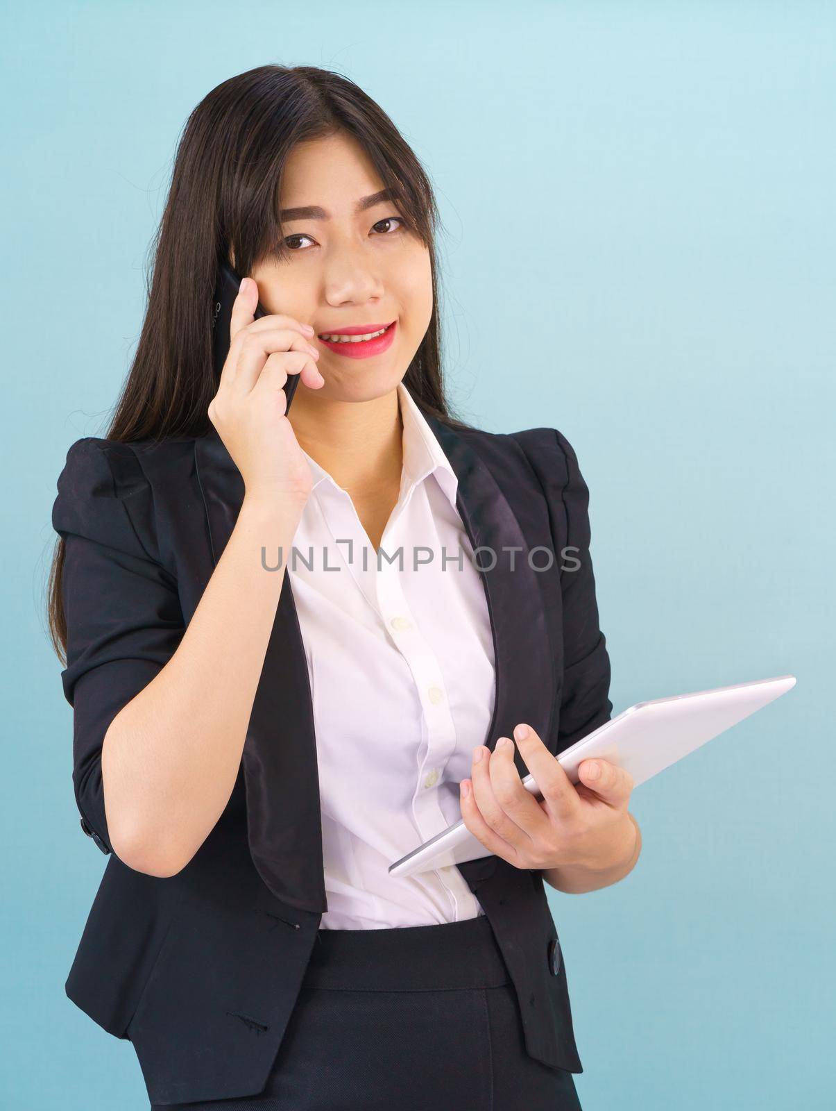 Young Asian women in suit standing using her phone and holding digital tablet against blue background