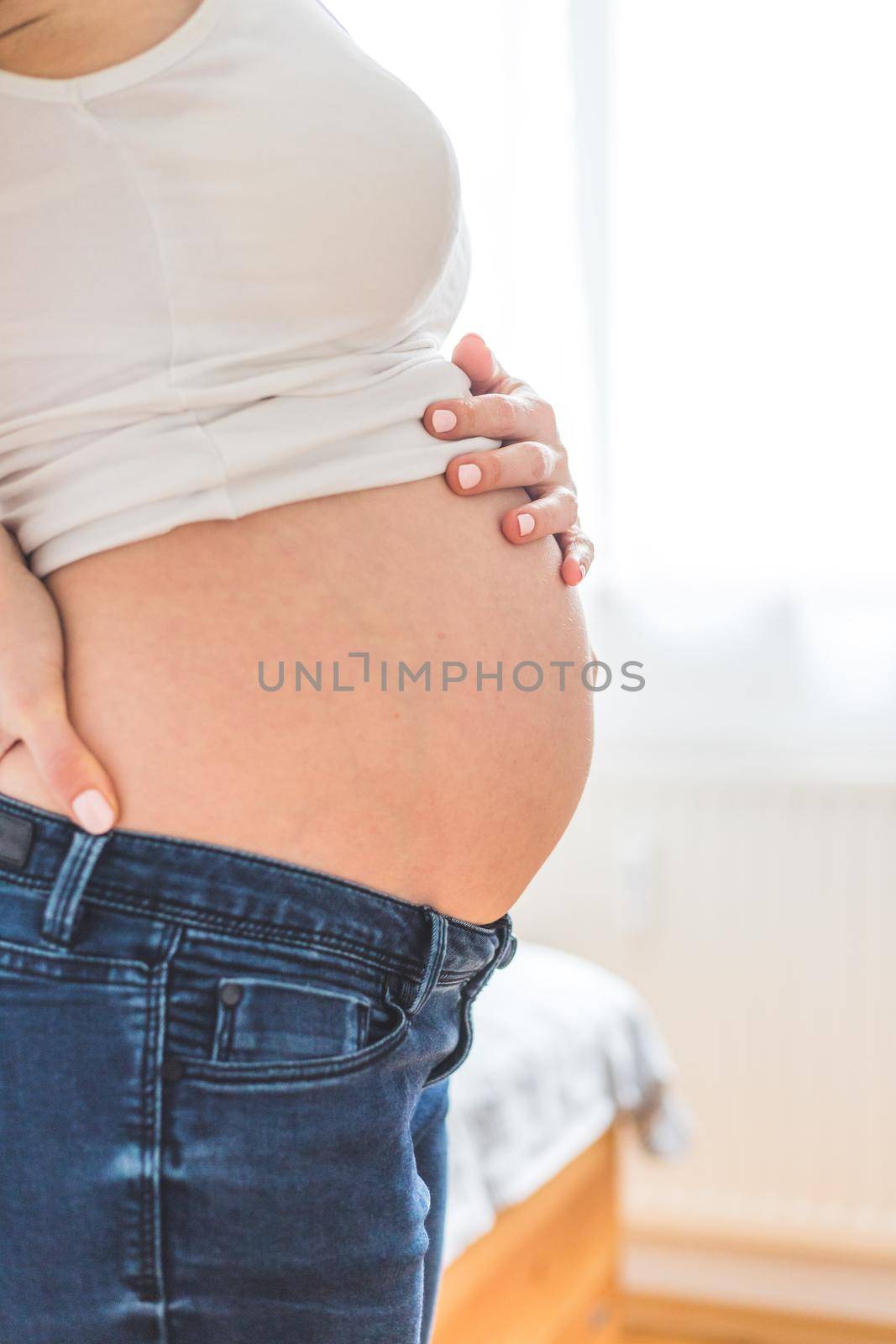 Happy pregnant in blue jeans mother is hugging her naked tummy, close up by Daxenbichler