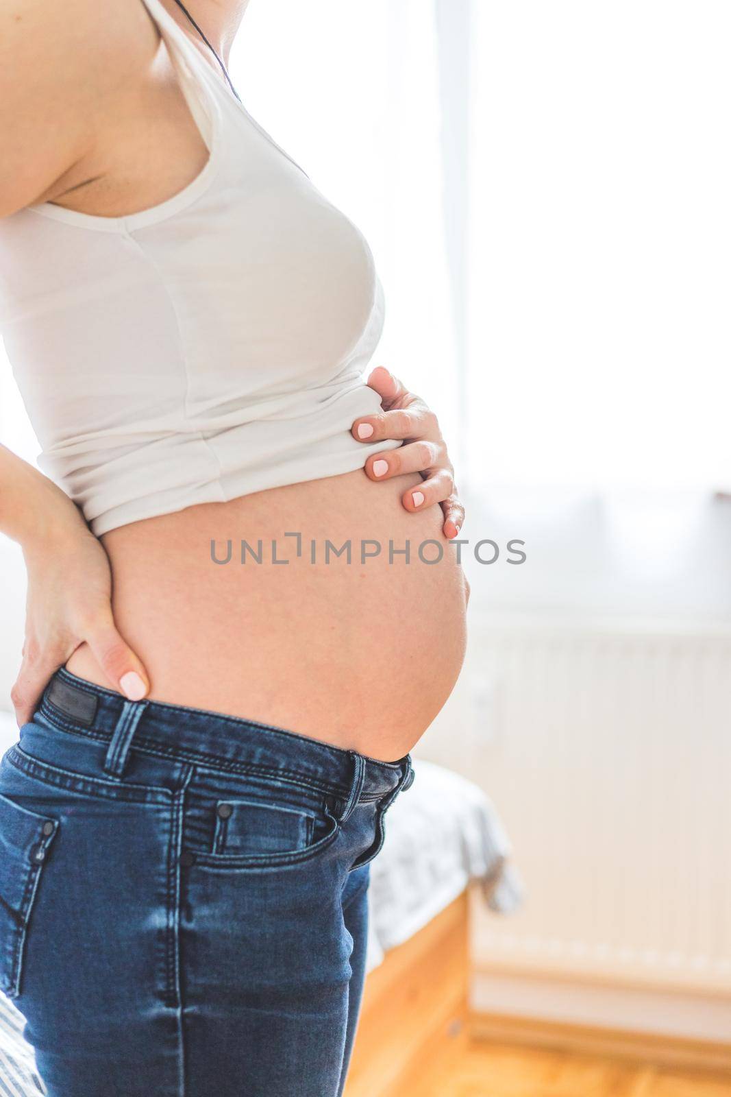 Happy pregnant in blue jeans mother is hugging her naked tummy, close up by Daxenbichler