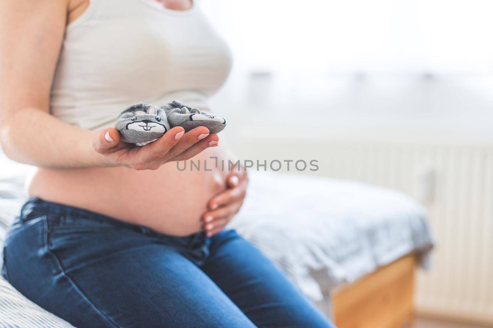 Caucasian pregnant mother is holding baby shoes in her hands, naked tummy in the blurry background