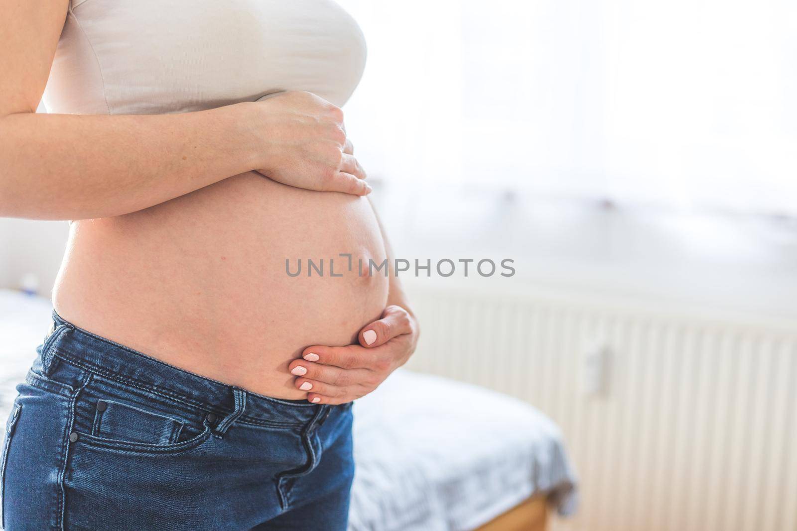 Happy pregnant in blue jeans mother is hugging her naked tummy, close up by Daxenbichler