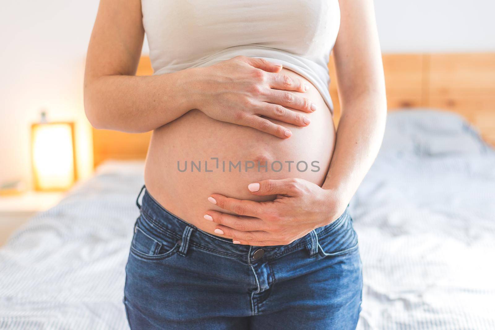 Happy pregnant in blue jeans mother is hugging her naked tummy, close up by Daxenbichler