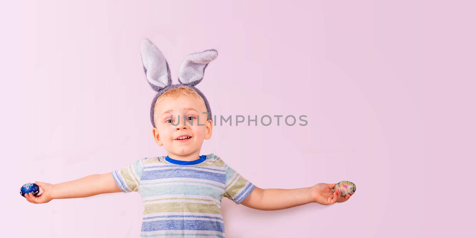 Cute baby boy in rabbit bunny ears on head and with colored eggs on pink background. Cheerful smiling happy child. Easter holiday banner