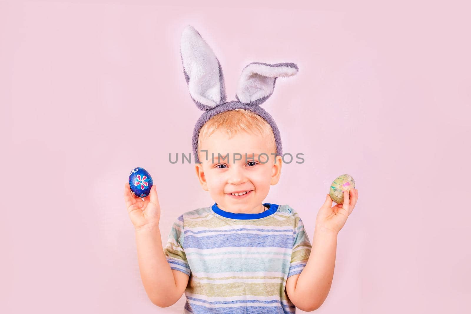 Cute baby boy in rabbit bunny ears on head and with colored eggs on pink background. Cheerful smiling happy child. Easter holiday