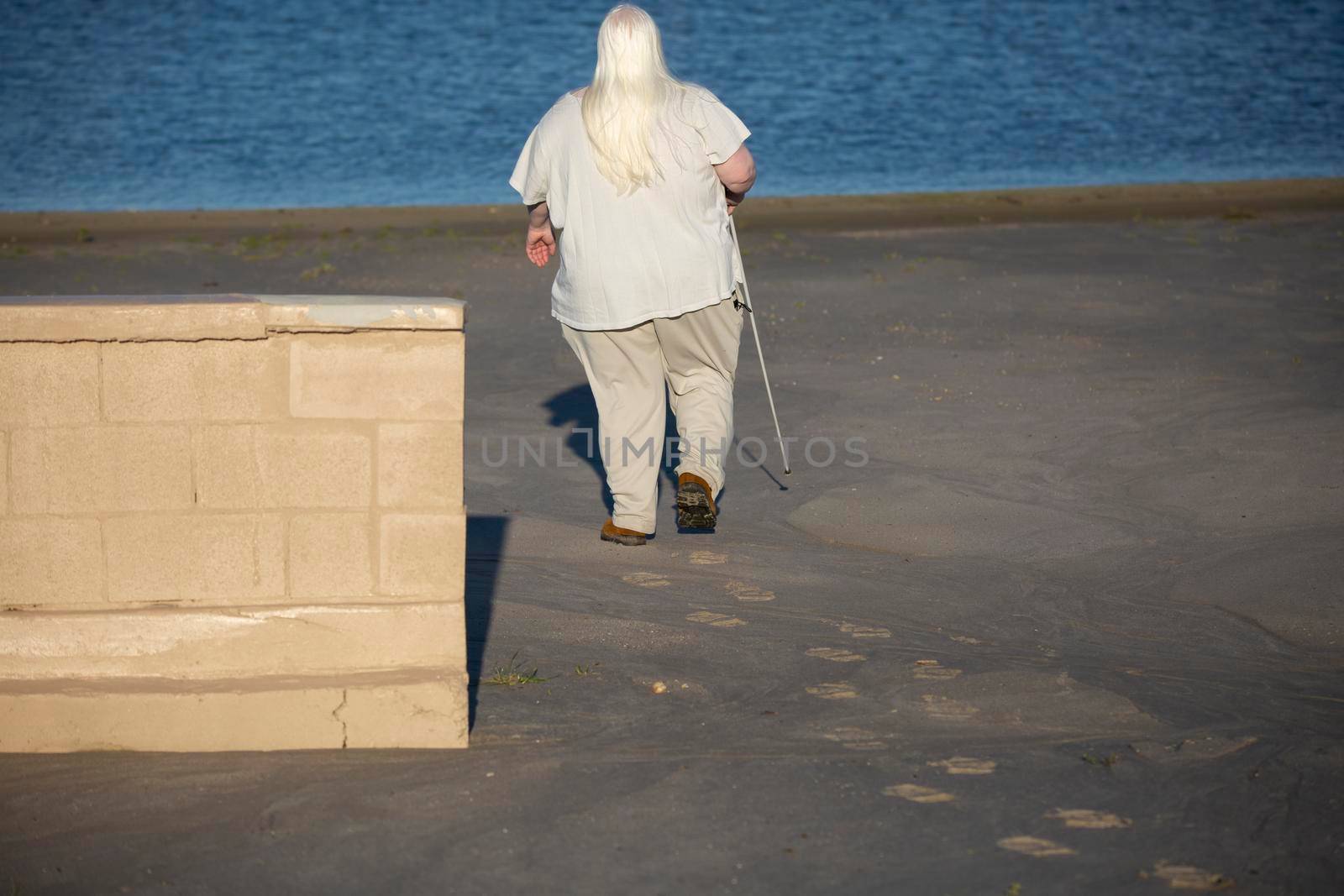 Blind woman walking past a tan brick wall toward water