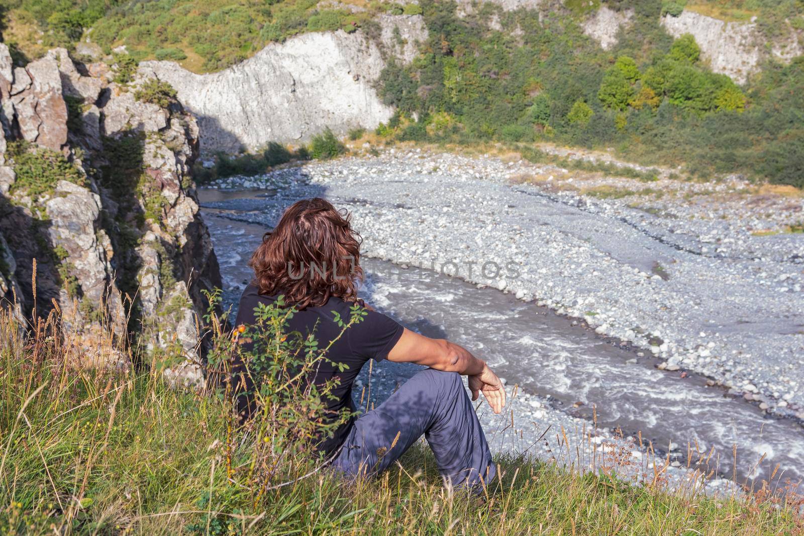 A man sits on a mountainside and looks at a stormy river by Yurich32