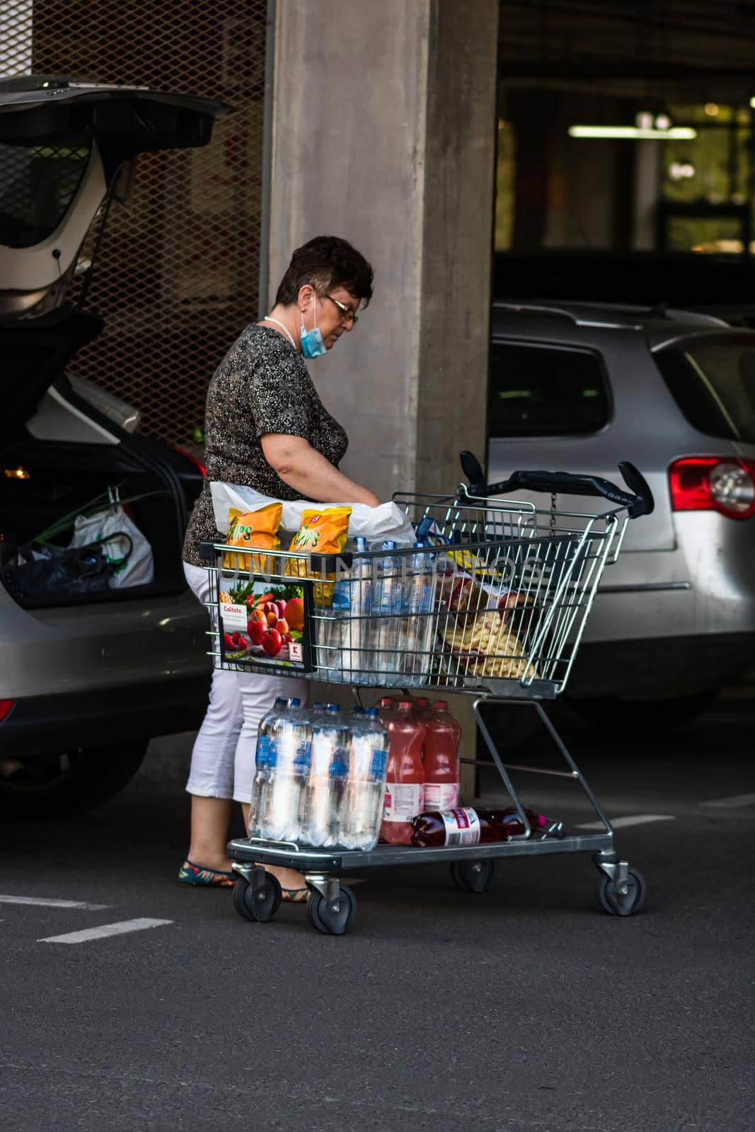 Woman wearing surgical mask with shopping cart full of goods, putting groceries in car trunk. Bucharest, Romania, 2020