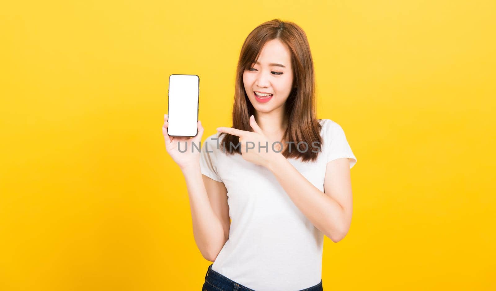 Asian happy portrait beautiful cute young woman smile standing wear t-shirt making finger pointing on smartphone blank screen looking to screen isolated, studio shot yellow background with copy space