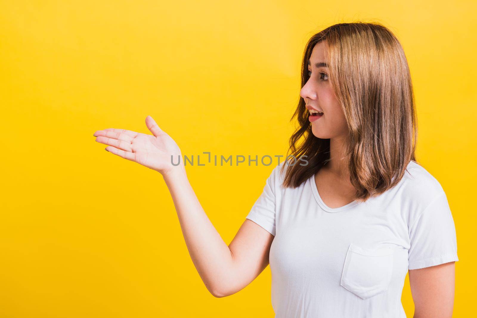 Asian Thai happy portrait beautiful cute young woman standing wear white t-shirt holding something on palm away side looking to side, studio shot isolated on yellow background with copy space