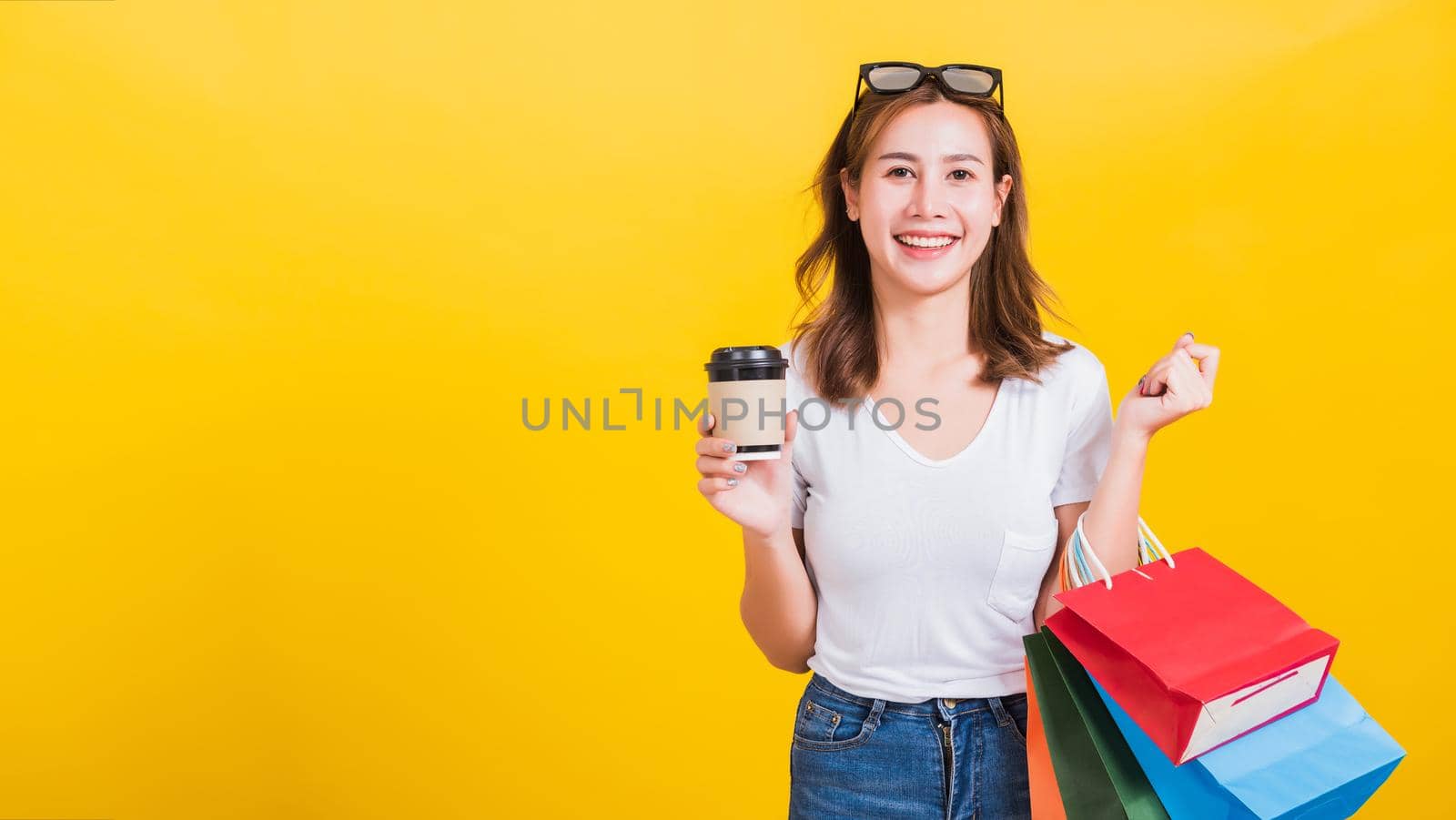 Portrait Asian Thai beautiful happy young woman smiling hold shopping bags multi-color and take away coffee cup her looking to camera, studio shot isolated on yellow background, with copy space