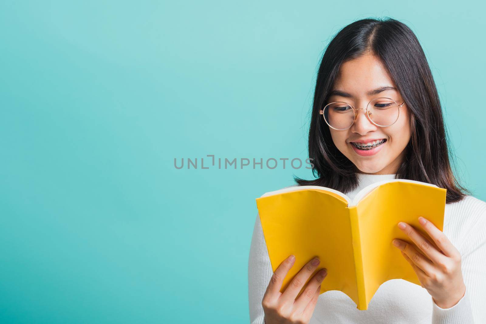 Portrait female in glasses is holding and reading a book, Young beautiful Asian woman hiding behind an open book, studio shot isolated on a blue background, Education concept