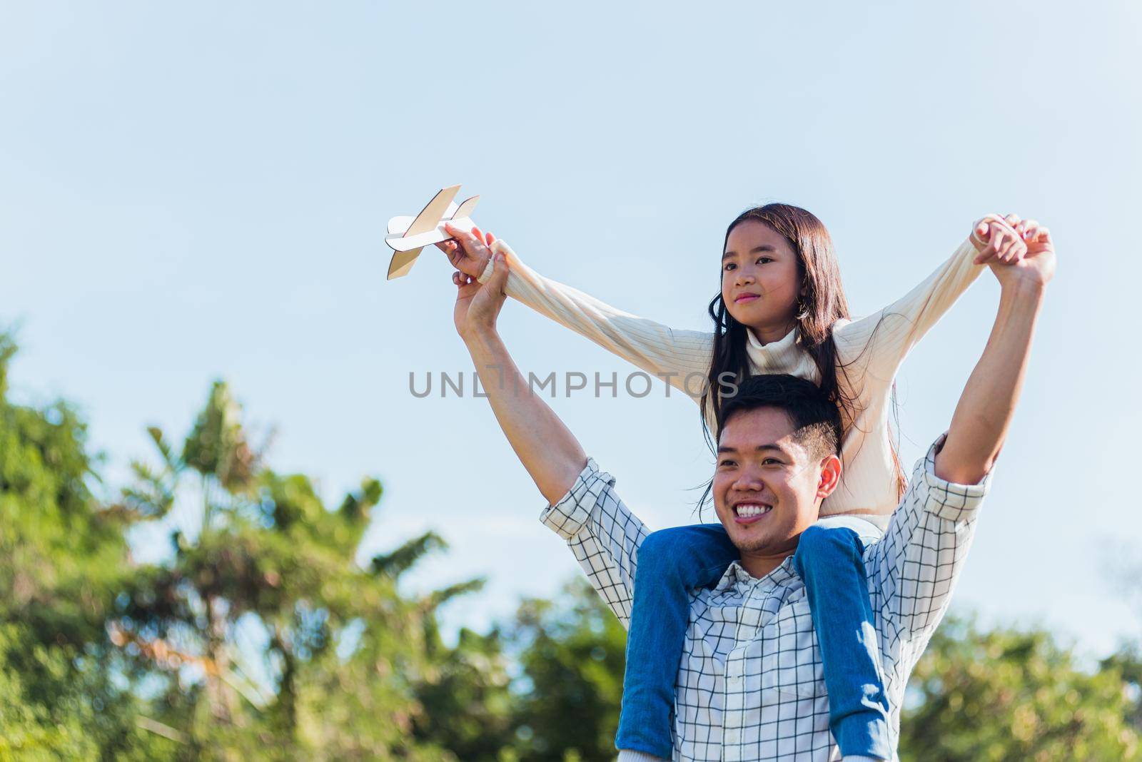 father and carrying an excited girl on shoulders having fun and enjoying outdoor lifestyle together playing aircraft toy by Sorapop
