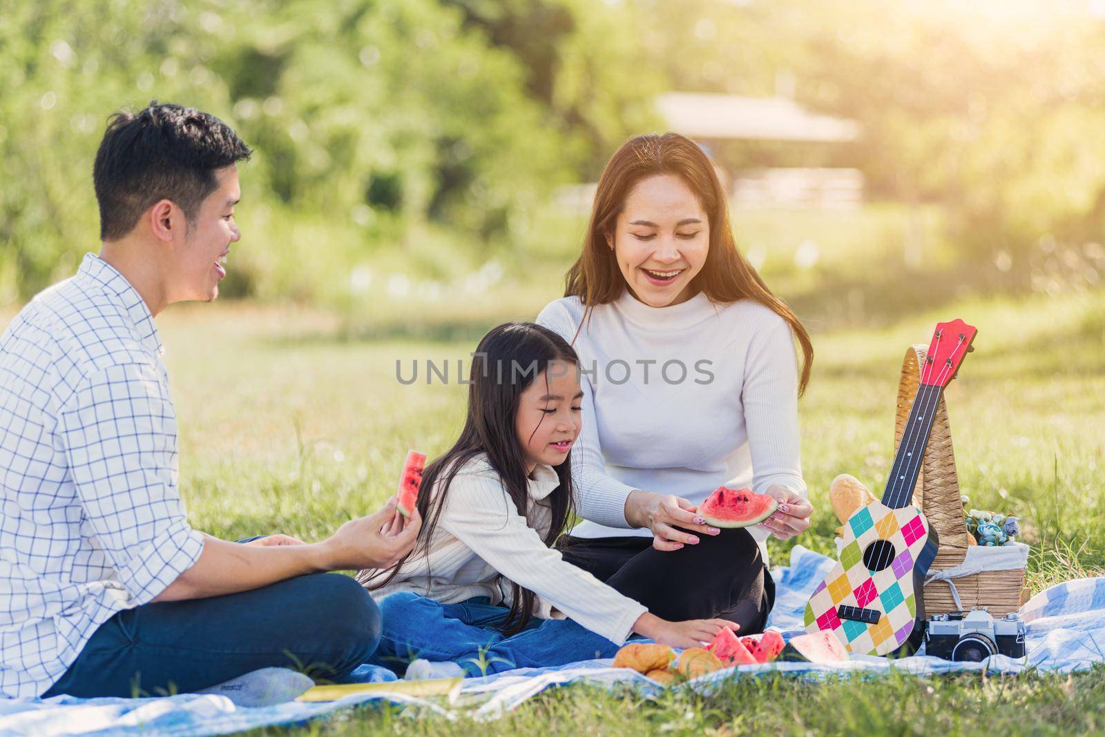Happy family having enjoying outdoor sitting on picnic blanket eating watermelon in park sunny time by Sorapop