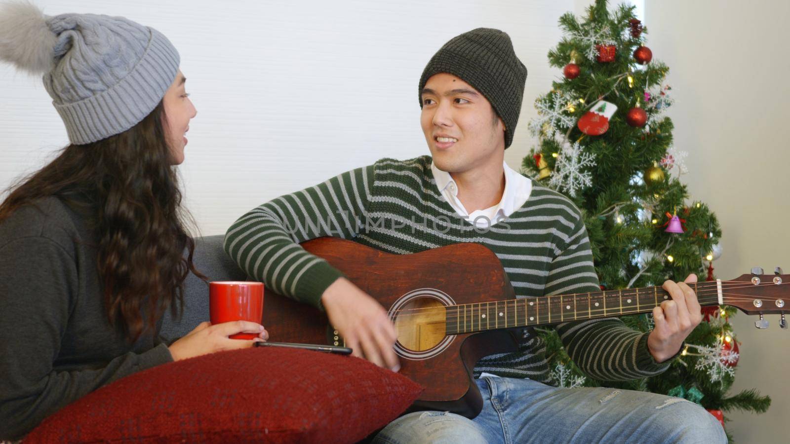 Happy Asian beautiful young family couple husband and wife on sofa great time in home living room, man and woman sit sofa playing guitar singing song winter holidays together enjoying,