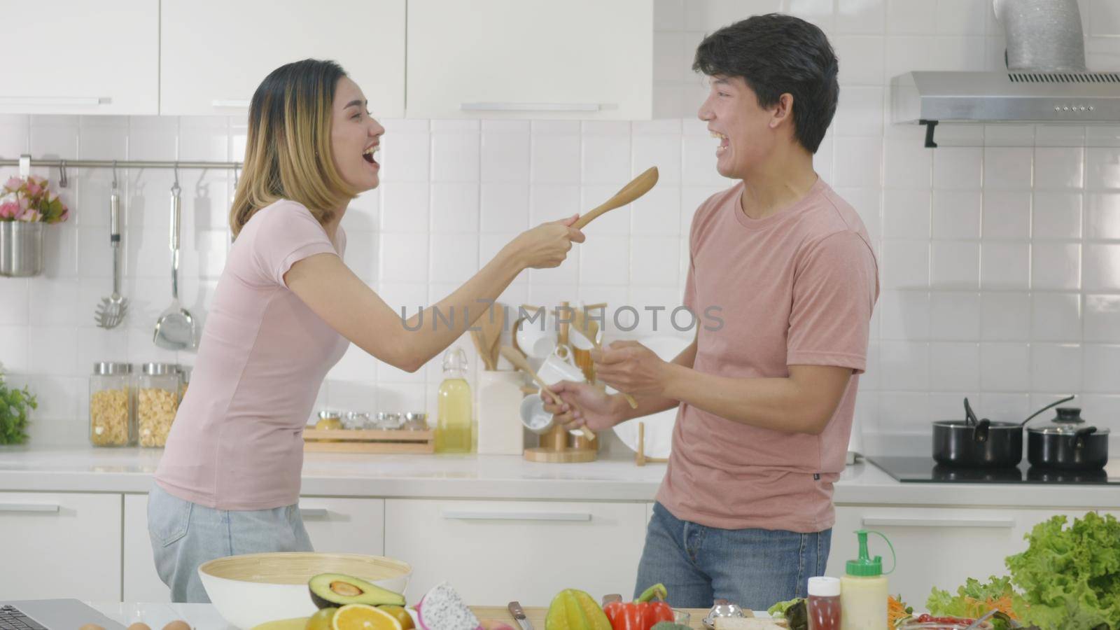 Happy Asian beautiful family couple husband and wife singing in kitchenware microphones in kitchen together having fun dance listen music at home. Two people dancing