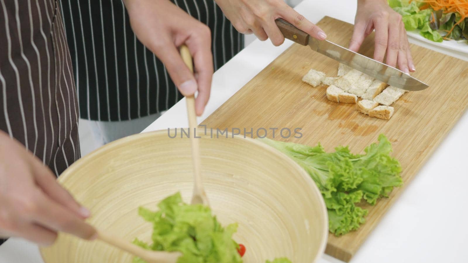 Happy Asian beautiful family couple husband and wife cooking vegetable salad in kitchen together at home. The man mix the vegetables in a cup woman sliced bread. healthy food concept