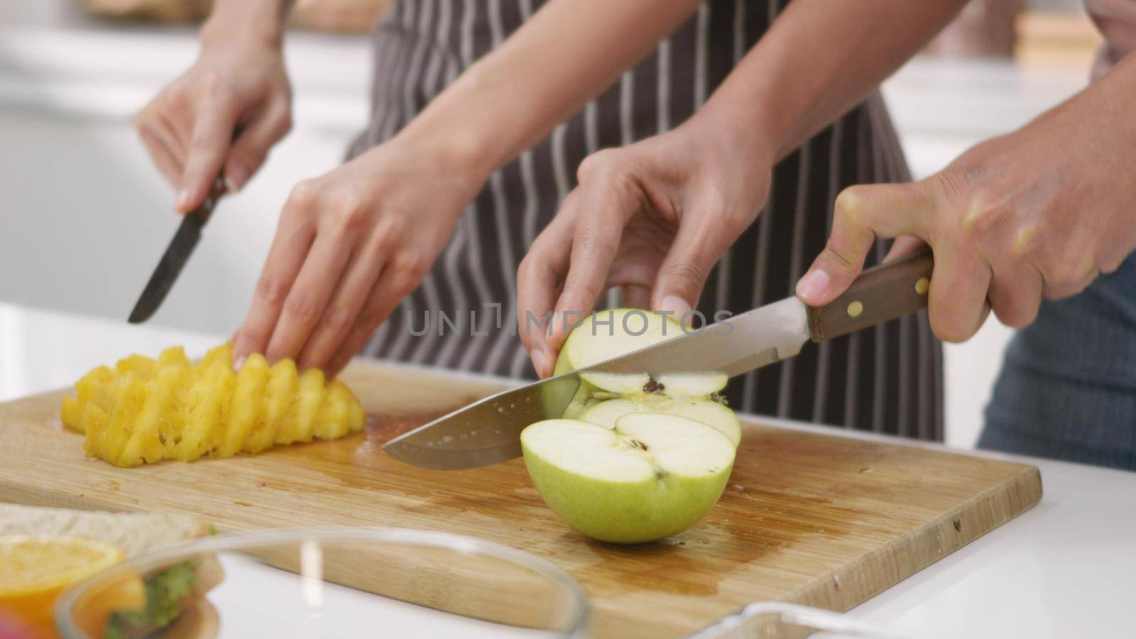 Happy Asian beautiful young family couple husband and wife making fresh smoothie in kitchen together at home. The man and woman slicing fresh apple fruits and pineapple. Healthy lifestyle concept