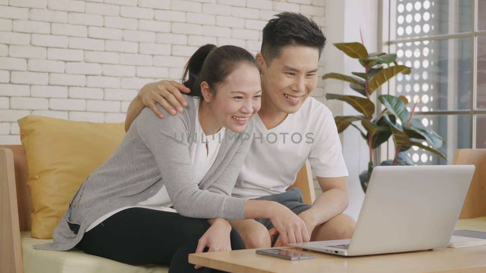 Happy Asian family couple husband and wife laughing sitting on sofa using laptop computer webcam technology talking making online social distance video call to their children at home.