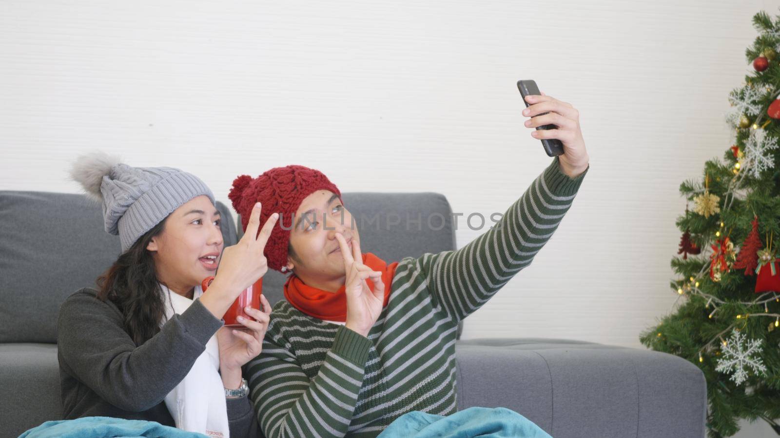 Happy Asian beautiful young family couple husband and wife sitting on sofa in living room, lovely woman and man funny making taking selfie together with smartphone,