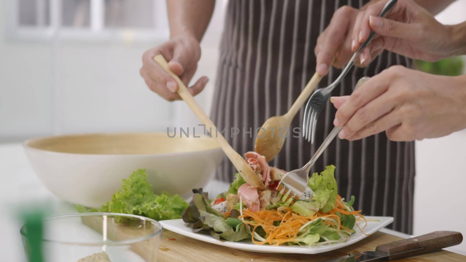 Happy Asian beautiful family couple husband and wife cooking vegetable salad in kitchen together at home. The man and woman help each other scoop salad vegetables on a plate. healthy food concept
