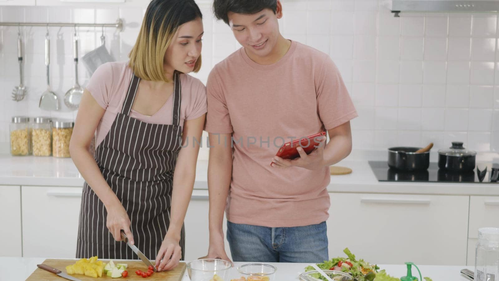 Happy Asian beautiful young family couple husband and wife cooking vegetable salad in kitchen together at home. The man and woman according online cooking class to recipe on laptop computer
