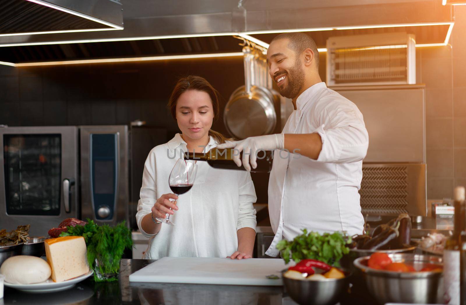 Handsome young African chef is cooking together with Caucasian girlfriend in the kitchen using red wine ingredient. A cook teaches a girl how to cook. Man and woman cooking in professional kitchen.