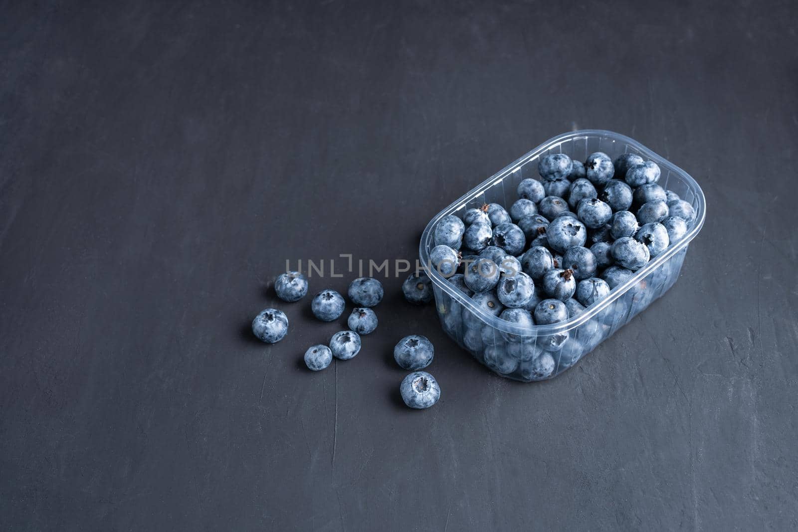 Tasty juicy raw blueberries in a plastic container on a black dark background. Packaging for berries in a supermarket on store shelves.