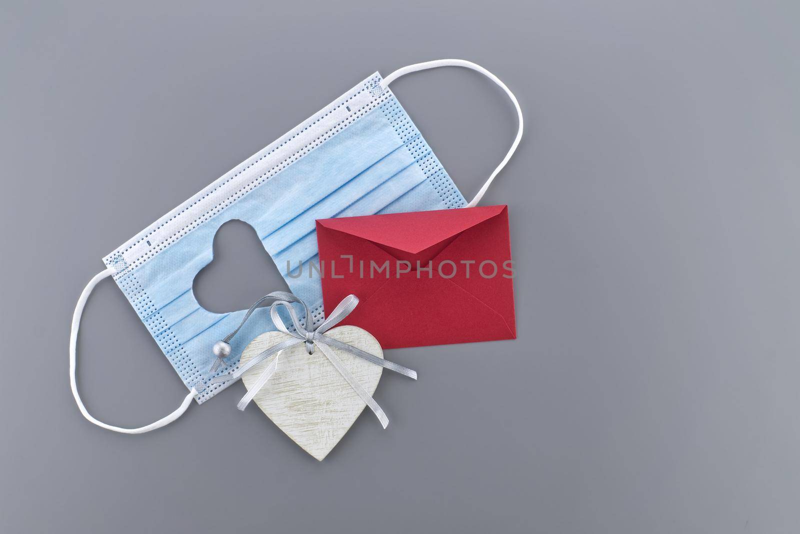 Medical mask with cut out heart shape, red rose and wooden heart medallion on gray background with free copy space