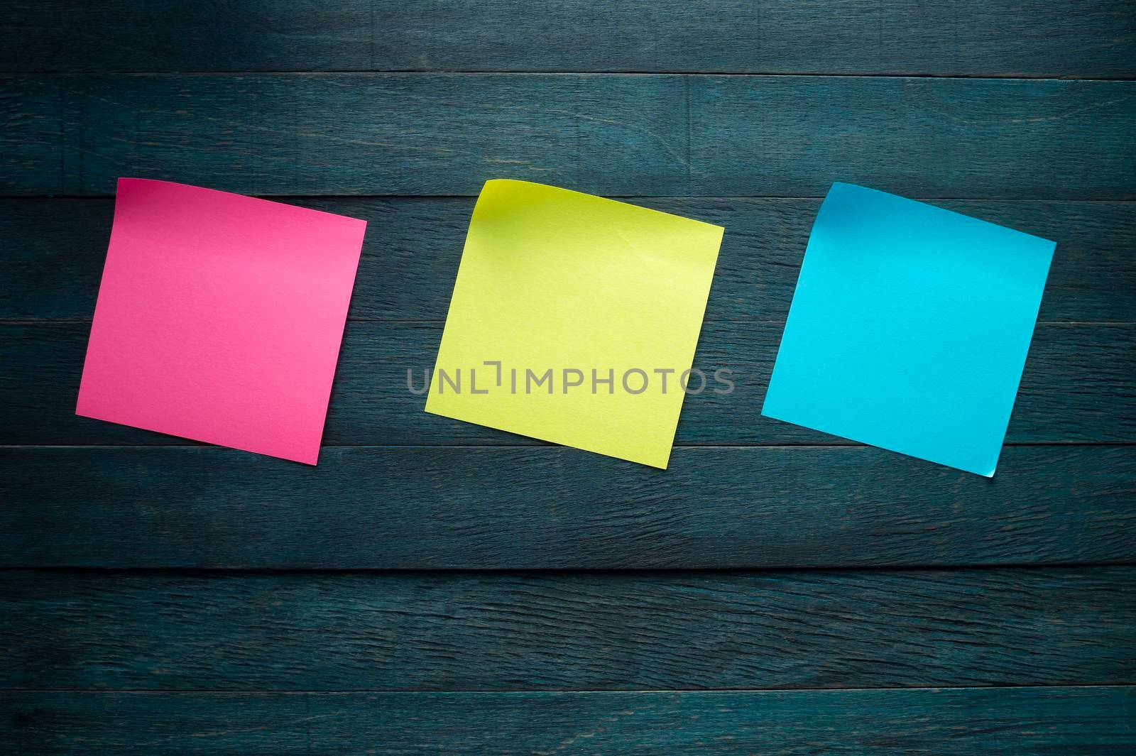 Pink yellow and blue blank adhesive note stickers with curled corners on blue rustic board. Free copy space for text