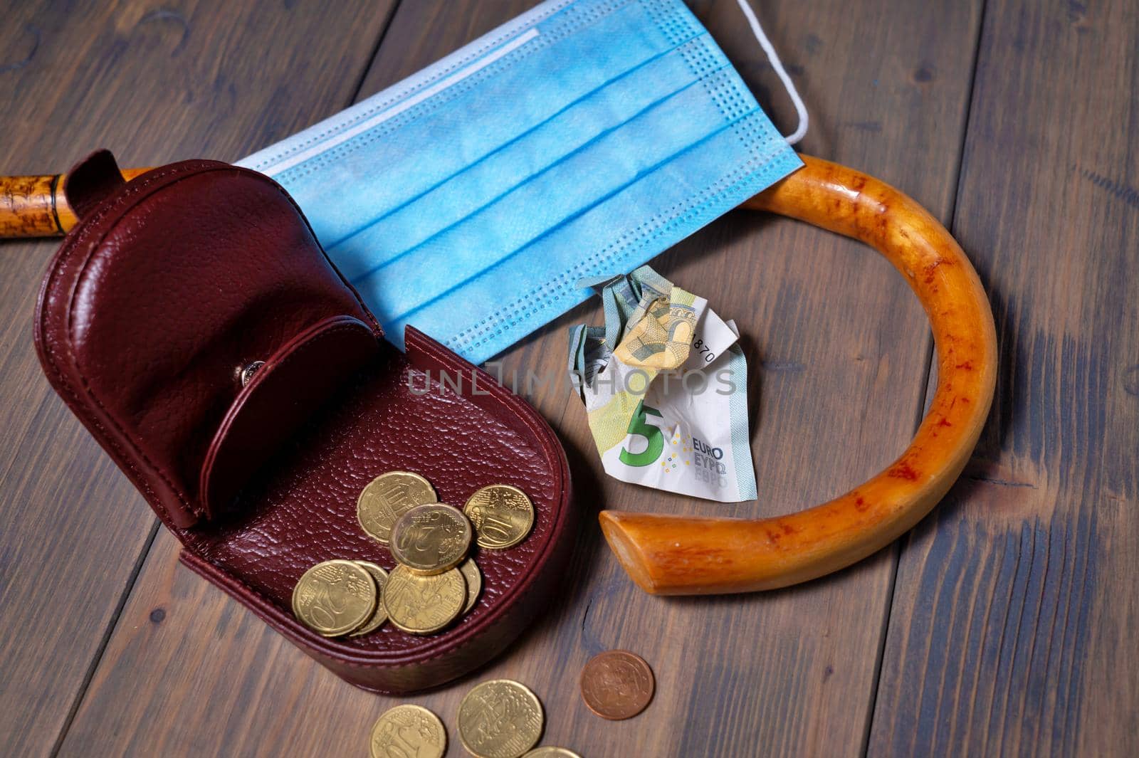 Conceptual Covid-19 flat lay still life with money spilling from an open purse onto a wooden table with cane and surgical mask alongside viewed from above