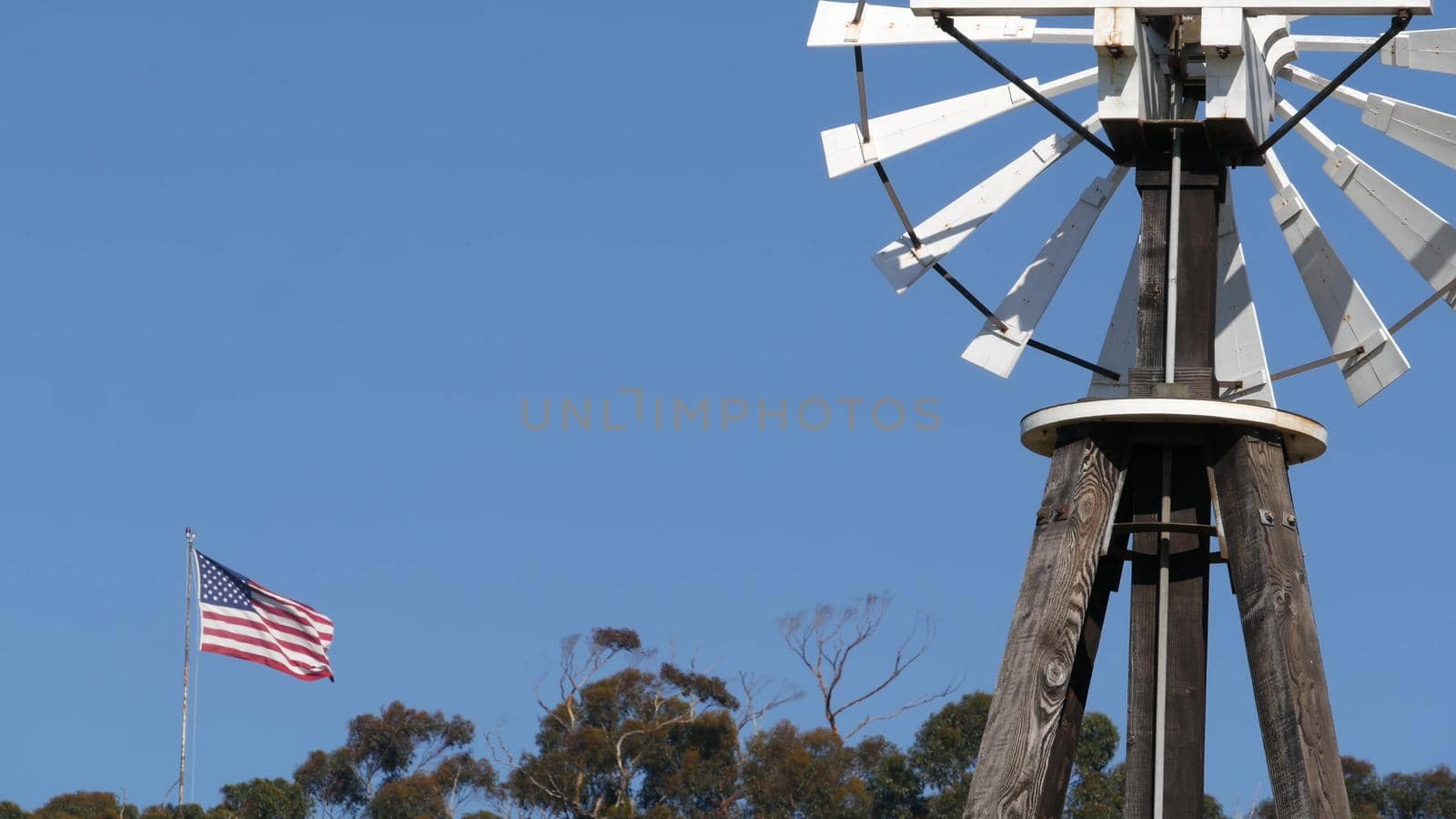 Classic retro windmill, bladed rotor and USA flag against blue sky. Vintage water pump wind turbine, power generator on livestock ranch or agricultural farm. Rural symbol of wild west, rustic suburb.