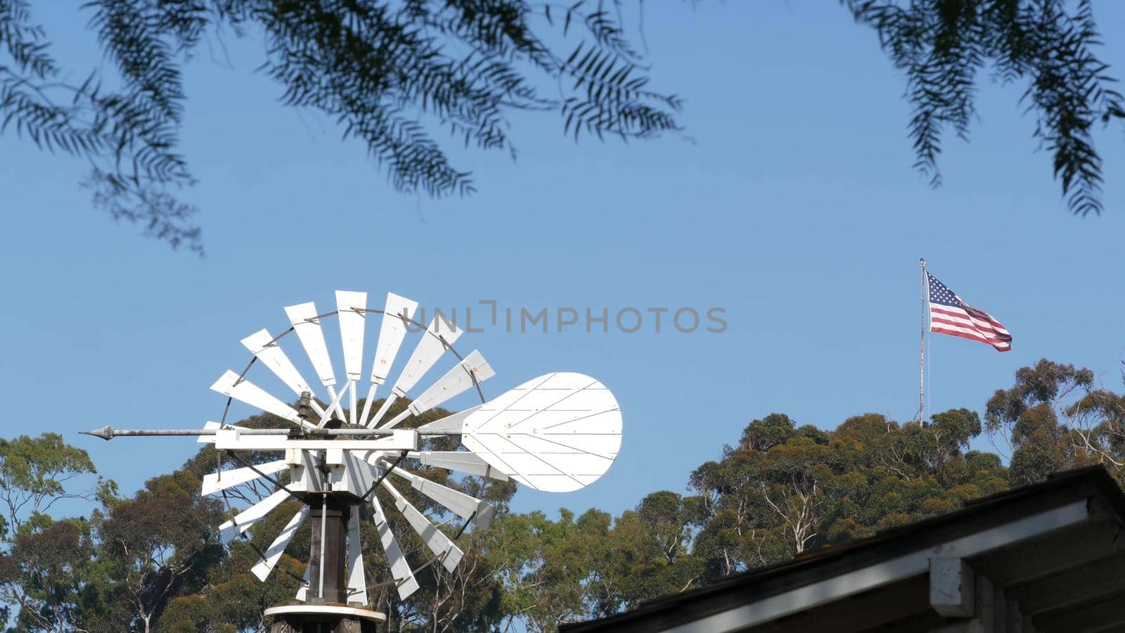 Classic retro windmill, bladed rotor and USA flag against blue sky. Vintage water pump wind turbine, power generator on livestock ranch or agricultural farm. Rural symbol of wild west, rustic suburb by DogoraSun