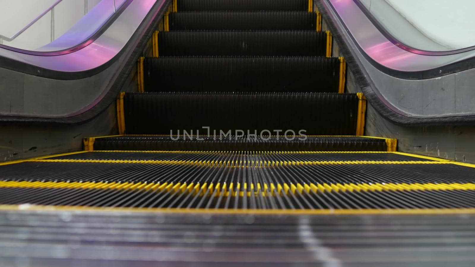 Low angle looped perspective view of modern escalator stairs. Automated elevator mechanism. Yellow line on stairway illuminated with purple light. Futuristic empty machinery staircase moving straight.