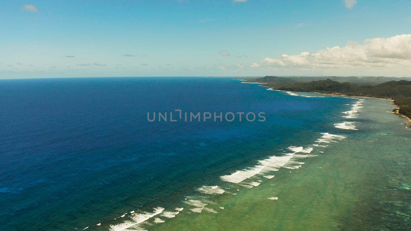 Coast of Siargao island is covered with forests and waves crashing on a coral reef. Seascape: ocean with blue water against the sky and clouds.