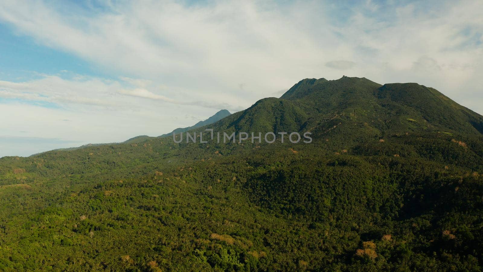 Mountains covered rainforest, trees and blue sky with clouds, aerial view. Camiguin, Philippines. Mountain landscape on tropical island with mountain peaks covered with forest. Slopes of mountains with evergreen vegetation.
