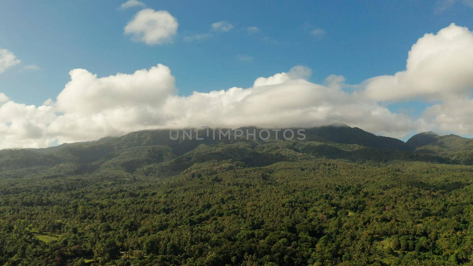 Mountain landscape on tropical island with mountain peaks covered with forest from above. Mountains covered rainforest, trees and blue sky with clouds, aerial view. Camiguin, Philippines. Slopes of mountains with evergreen vegetation.