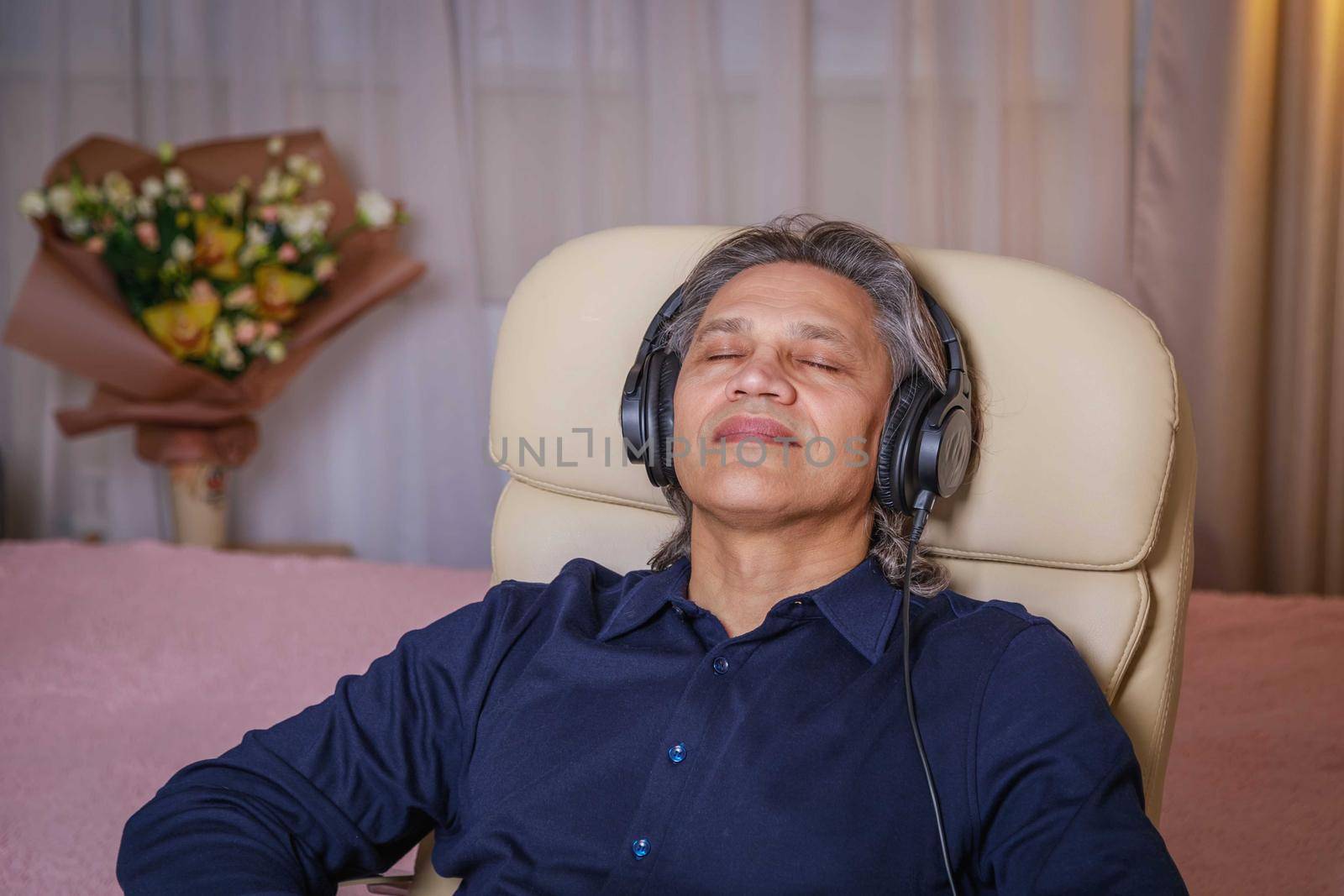 50-year-old man listens to music on headphones at home, sitting in a chair. by Yurich32