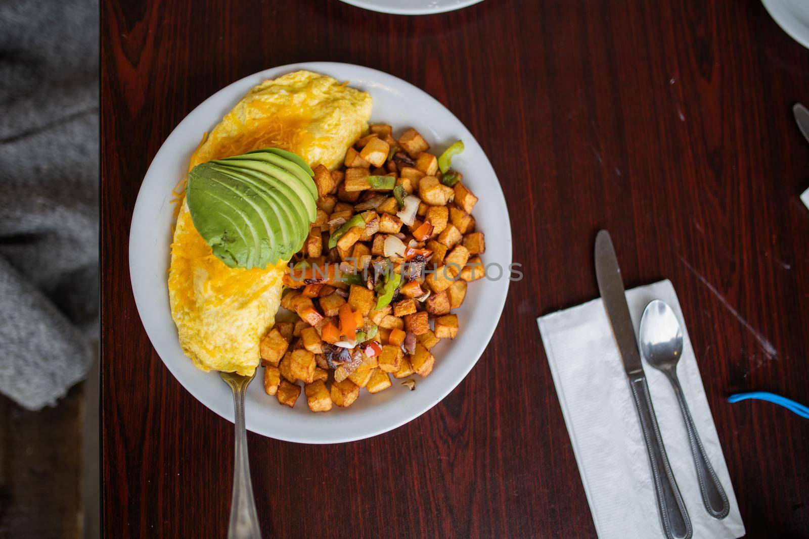 Top view of omelette, roasted potatoes and avocado slices on white plate above wooden table. Cutlery and tasty-looking dish with eggs and chopped potatoes. Delicious balanced diet