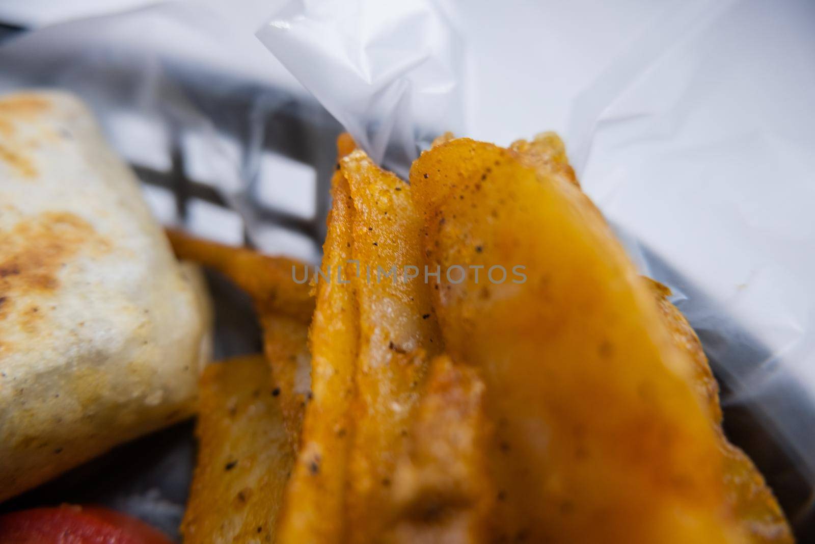 Extreme close-up of potato wedges and two spicy pork burritos in black plastic basket. Mexican-style pork burritos and vegetables in plastic container. Mexican fast food