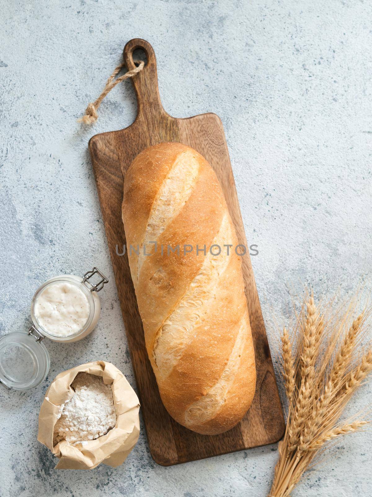 British White Bloomer or European sourdough Baton loaf bread on gray cement background. Fresh loaf bread, glass jar with sourdough starter, flour in paper bag and ears. Top view. Copy space. Vertical