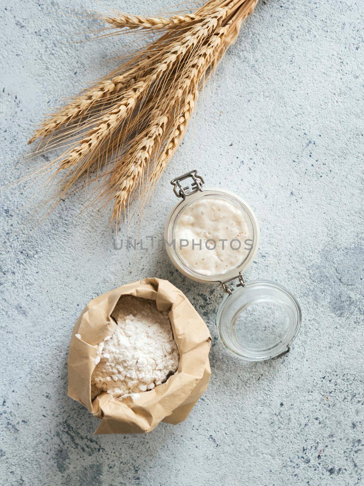 Wheat sourdough starter. Top view of bread making ingredients - glass jar with sourdough starter, flour in paper bag and ears over gray cement background. Copy space for text or design. Vertical.