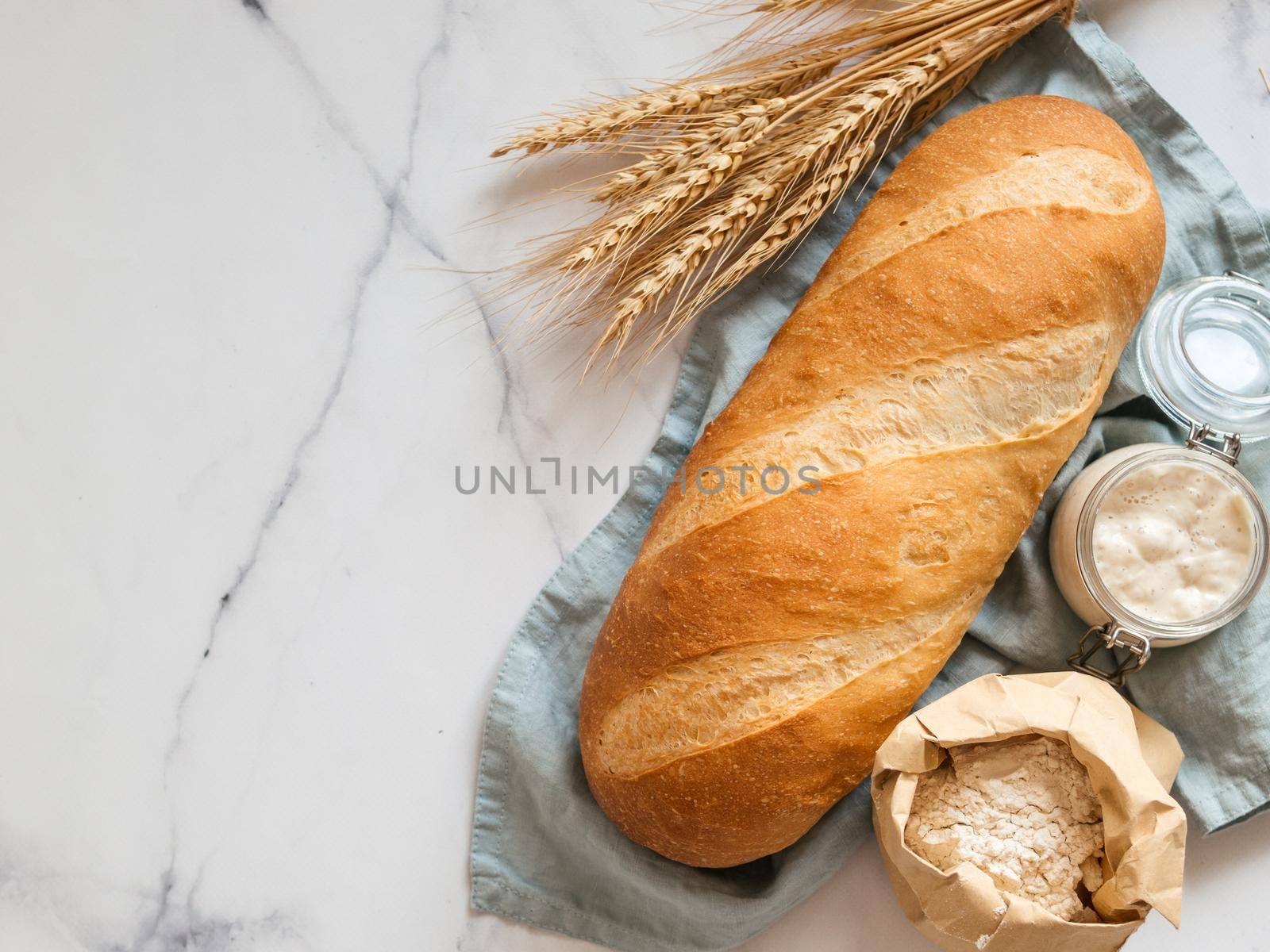 British White Bloomer or European sourdough Baton loaf bread on white marble background. Fresh loaf bread and glass jar with sourdough starter, flour in paper bag and ears. Top view. Copy space