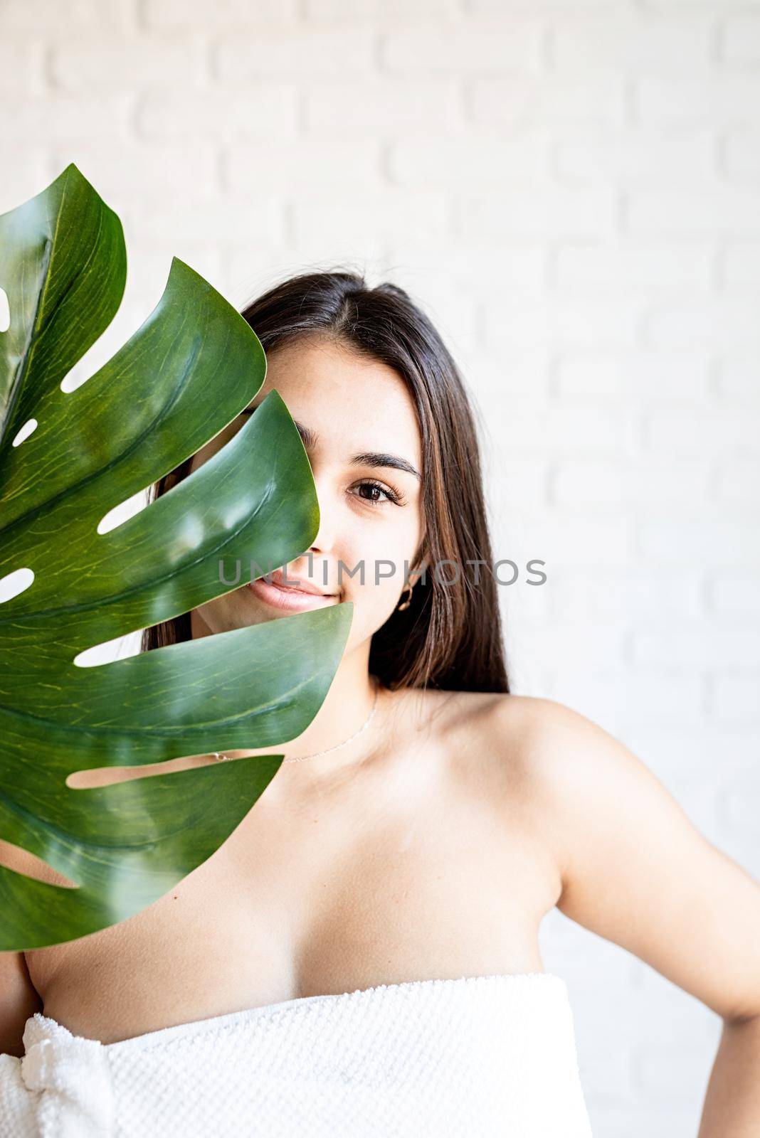 Spa Facial Mask. Spa and beauty. Happy beautiful brunette woman wearing bath towels holding a green monstera leaf in front of her face